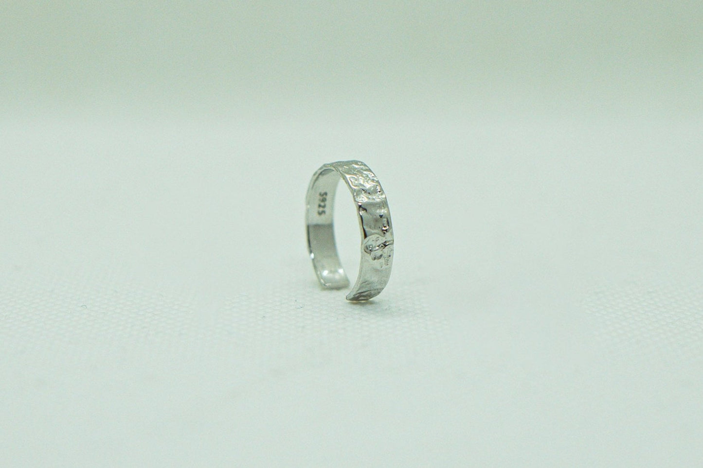 Einzelner, offener Silberring mit gehämmert wirkender Textur, präsentiert auf einem hellen, einfarbigen Hintergrund. Der Ring, mit seiner rustikalen Eleganz und dem einzigartigen Design, ist das zentrale Element des Bildes. Silberring, gehämmert, offener Ring, unisexring, Schmuck, Modeschmuck, Accessoires, handgefertigter Schmuck.