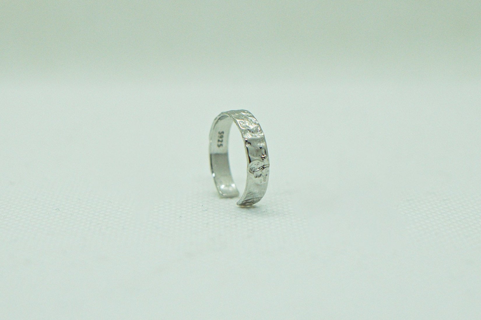 Einzelner, offener Silberring mit gehämmert wirkender Textur, präsentiert auf einem hellen, einfarbigen Hintergrund. Der Ring, mit seiner rustikalen Eleganz und dem einzigartigen Design, ist das zentrale Element des Bildes. Silberring, gehämmert, offener Ring, unisexring, Schmuck, Modeschmuck, Accessoires, handgefertigter Schmuck.