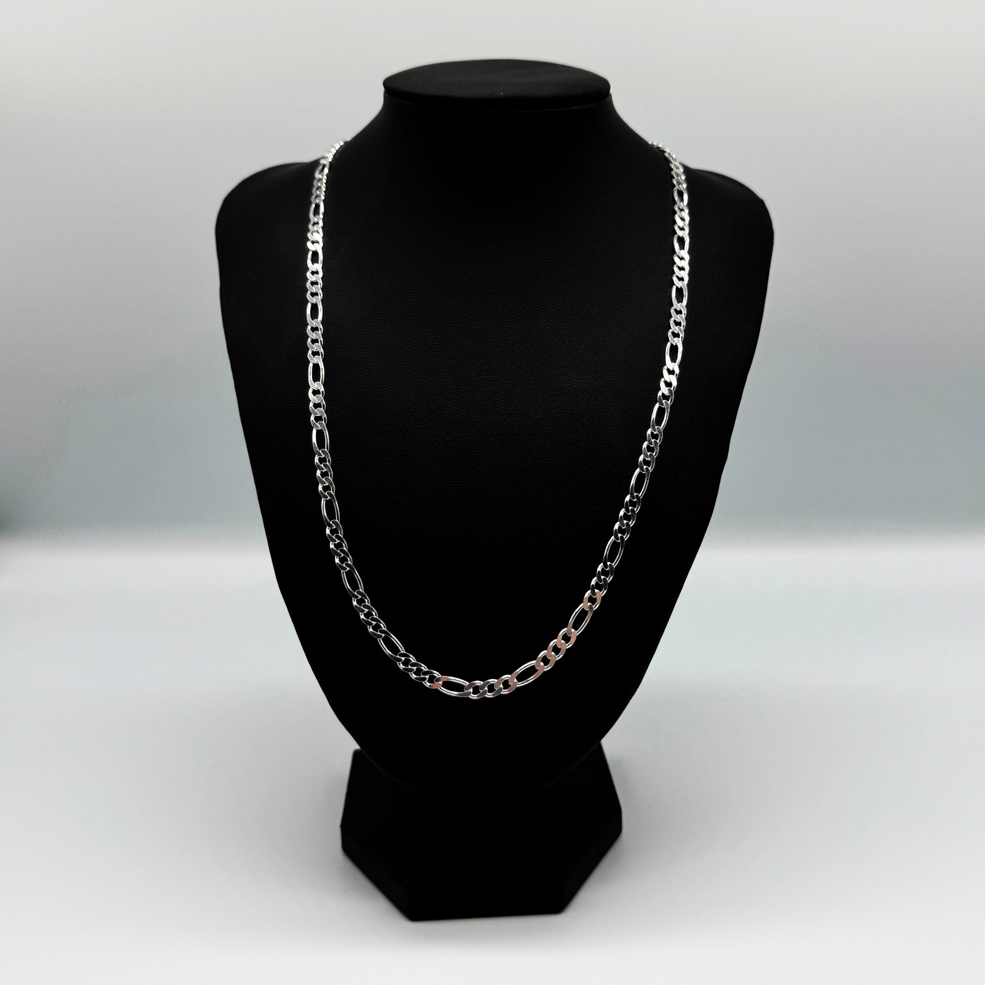 Eine silberne Figaro-Kettenhalskette auf einem schwarzen Lederständer vor einem weißen Hintergrund. Die Kette hat ein klassisches Design mit abwechselnd großen und kleinen Gliedern, ideal für den Alltag oder als elegantes Accessoire.