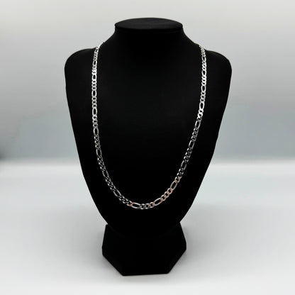 Eine silberne Figaro-Kettenhalskette auf einem schwarzen Lederständer vor einem weißen Hintergrund. Die Kette hat ein klassisches Design mit abwechselnd großen und kleinen Gliedern, ideal für den Alltag oder als elegantes Accessoire.