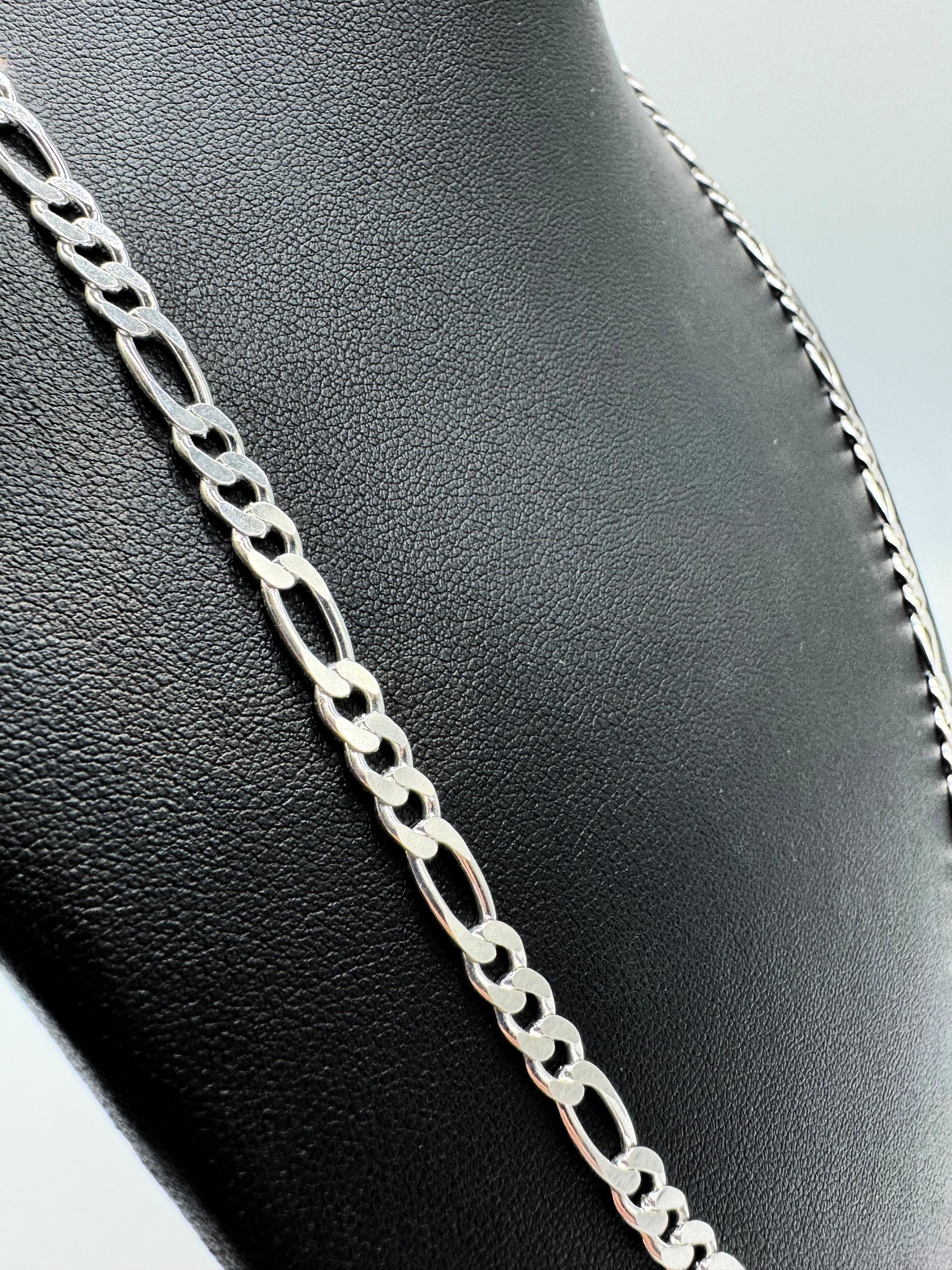 Eine silberne Figaro-Kettenhalskette auf einem schwarzen Lederständer vor einem weißen Hintergrund. Die Kette hat ein klassisches Design mit abwechselnd großen und kleinen Gliedern, ideal für den Alltag oder als elegantes Accessoire.